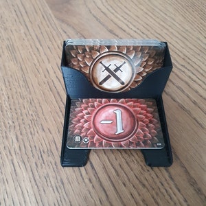 Puede incluir: Un porta tarjetas de plástico negro con dos tarjetas en su interior. La tarjeta superior tiene un fondo marrón con un símbolo de espadas cruzadas. La tarjeta inferior tiene un fondo rojo con un número blanco "1" dentro de un círculo.