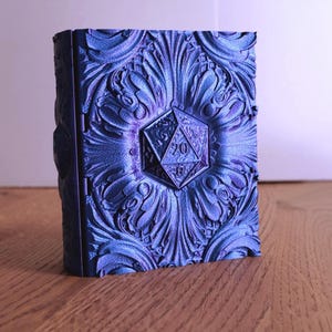May include: A blue, ornate box with a raised floral design and a 20-sided die emblem. The box has a hinged closure and is likely used for storing dice or small items. The colour is a metallic blue.