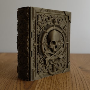 May include: An olive-green decorative book with a skull and crossbones motif on the front cover. The book features detailed, carved patterns and stands upright on a wooden surface. The spine and edges of the book are also intricately carved.