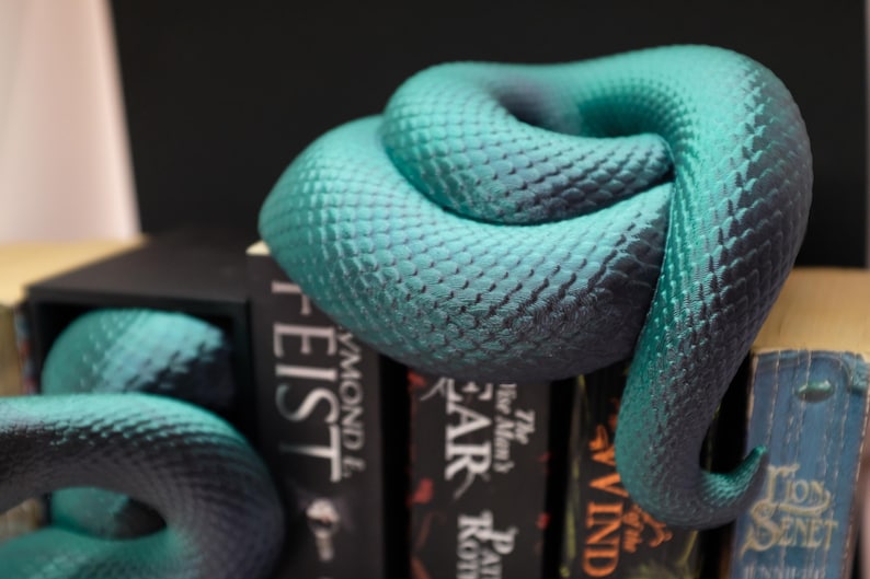 Puede incluir: Dos sujetalibros en forma de serpiente azul turquesa con una superficie texturizada, sosteniendo una pila de libros con t&iacute;tulos como "The Man's" y "Feist".