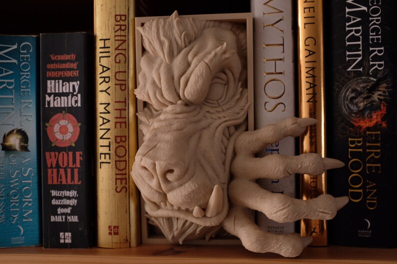 May include: A bookend featuring a sculpted monster with a snarling expression and a clawed hand. The bookend is made of a light brown material and is holding up a row of books. The books include titles like "Bring Up the Bodies" and "Fire and Blood".