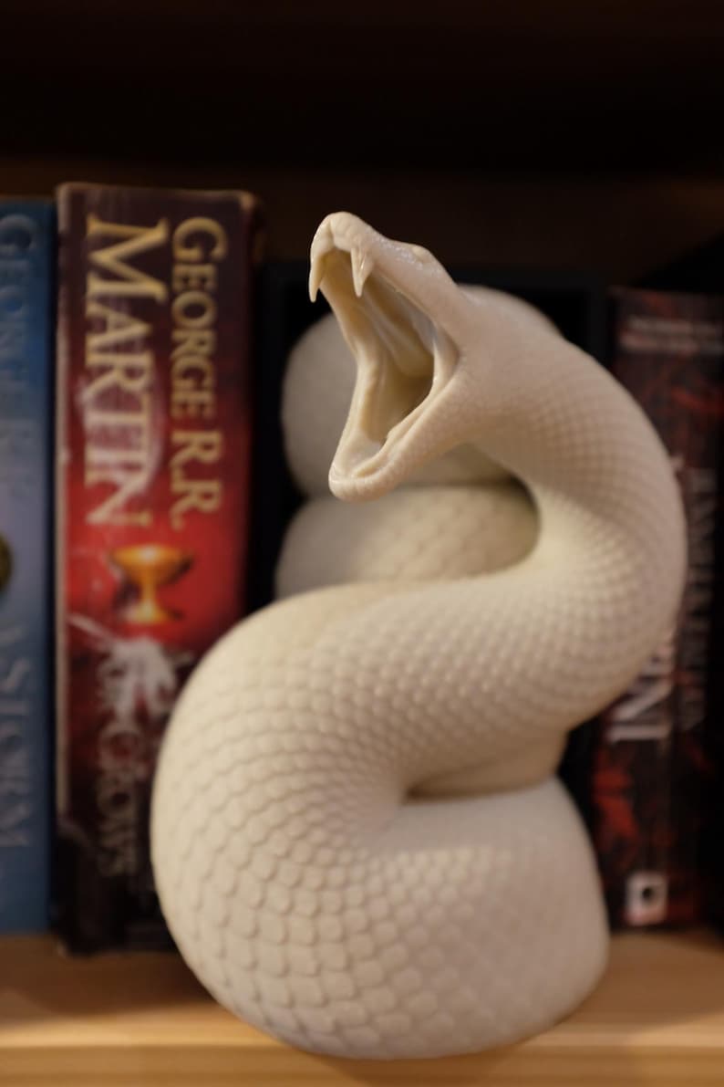 Puede incluir: Una escultura de serpiente de cer&aacute;mica blanca con la boca abierta, colocada en una estanter&iacute;a entre dos libros. La serpiente est&aacute; enroscada y tiene una superficie texturizada.