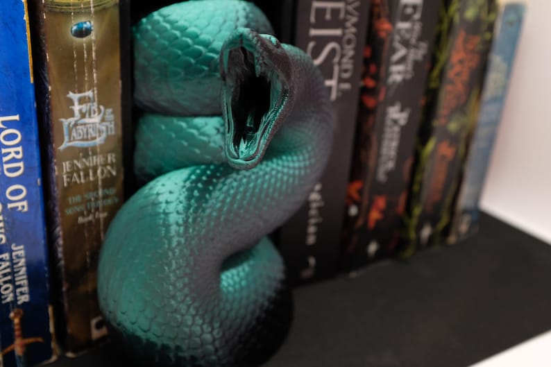 Puede incluir: Un sujetalibros de serpiente verde azulado con una superficie texturizada y la boca abierta. El sujetalibros sostiene una fila de libros.