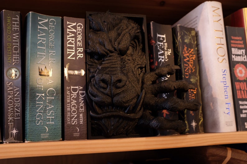 May include: A black dragon bookend with a textured surface, holding up a row of fantasy novels, including "A Clash of Kings" and "A Dance with Dragons" by George R.R. Martin, "The Witcher" by Andrzej Sapkowski, and "Mythos" by Stephen Fry.