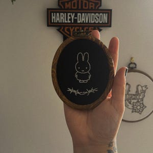 May include: Oval embroidery hoop with a white bunny design stitched on a black fabric. The wooden frame has a natural finish. The design includes stylized text at the bottom. A Harley-Davidson sign is in the background.