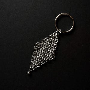May include: A silver chainmail keychain with a diamond-shaped pattern. The keychain is constructed from small, interlocking metal rings and has a small, round bead at the base. A circular key ring is attached at the top.