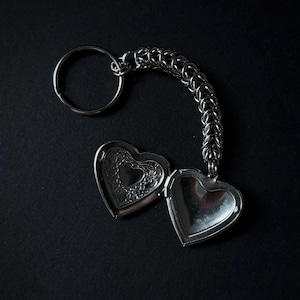 May include: Silver heart-shaped locket keyring, open to show the inside. The locket is attached to a chainmail-style loop and a keyring. The locket's interior has a heart design. The background is black.