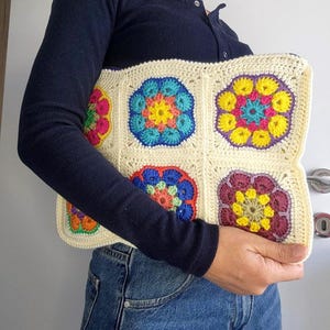 Könnte beinhalten: Cremefarbene Häkeltasche mit sechs bunten Blumenquadraten. Jedes Quadrat weist eine andere Farbkombination auf, darunter Rosa, Blau, Gelb und Lila. Die Tasche wird von einer Person gehalten, die ein marineblaues Langarmshirt und Jeans trägt.