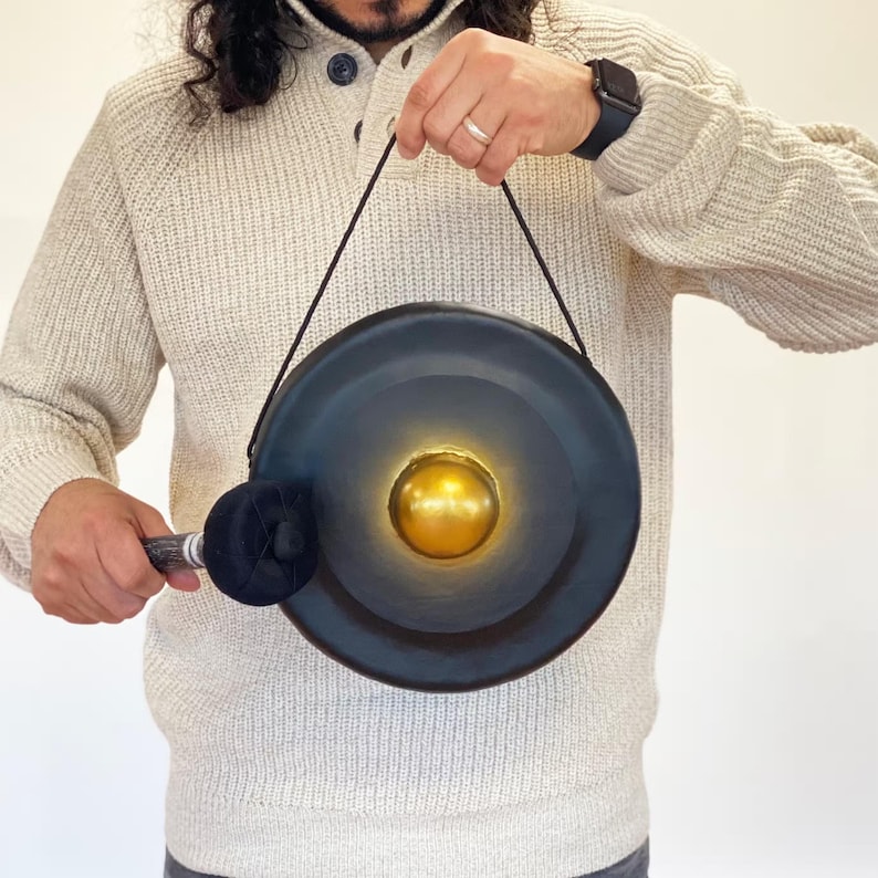 May include: A dark blue gong with a gold center, suspended by a black cord. A black mallet with a patterned handle rests against the gong. The gong is held by a person wearing a cream-colored sweater.