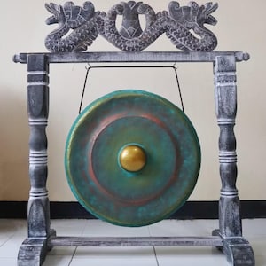 May include: A decorative gong with a teal and copper-colored face and a gold center. The gong hangs from a wooden frame with carved dragon accents. The frame is painted in shades of gray and white.
