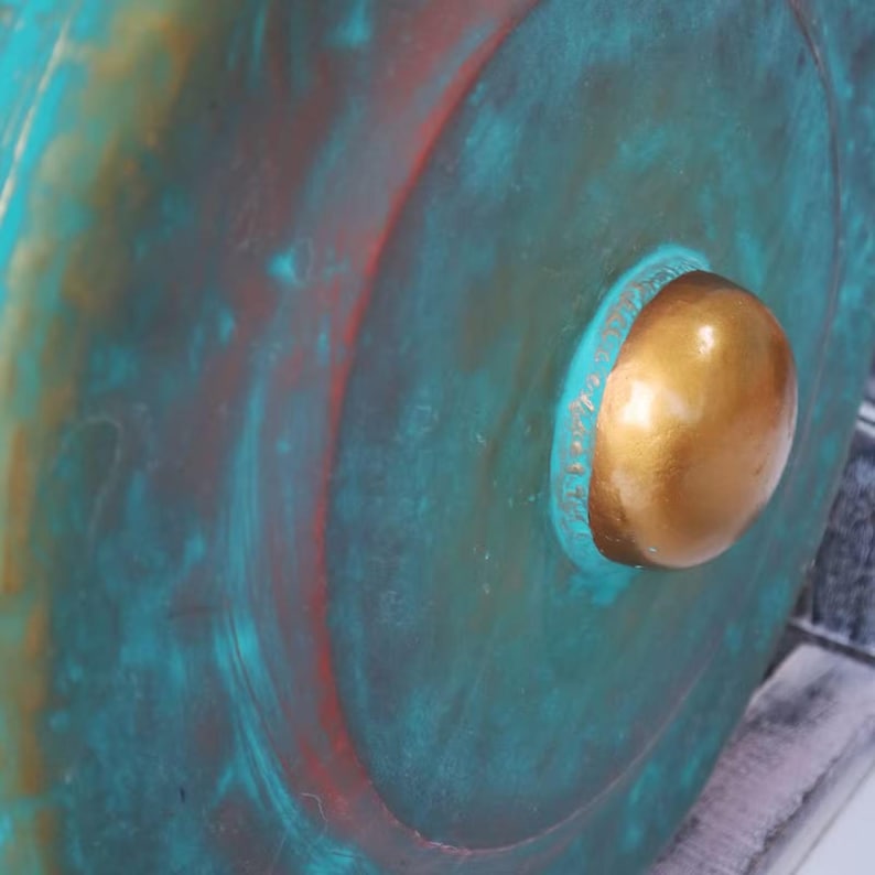 May include: Close-up of a weathered, circular object with a prominent, gold-colored knob. The surface has a distressed, aged appearance with teal, red, and gold tones, suggesting a vintage or antique aesthetic. The object's texture appears rough.