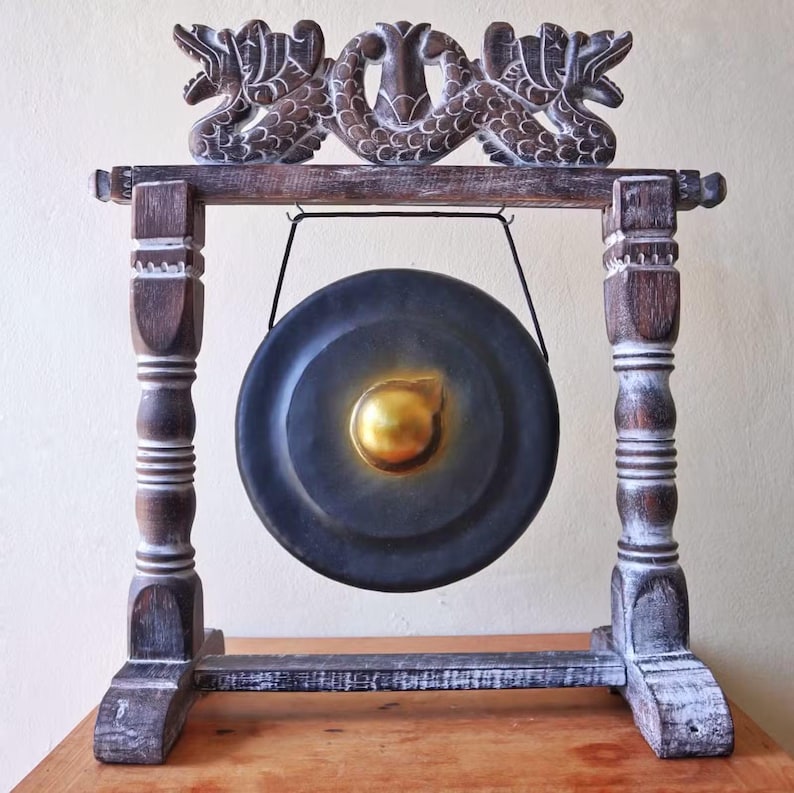May include: A decorative gong with a dark metal disc and a gold center, suspended within a wooden frame. The frame features carved dragon-like figures and ornate, distressed pillars. The gong is displayed on a wooden surface.