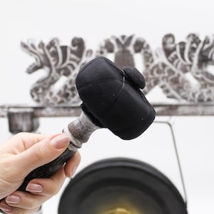 May include: A black mallet with a rounded head and a wooden handle. The mallet is held in a hand, with a decorative carved wooden structure in the background. The mallet is likely used for striking a gong or similar instrument.