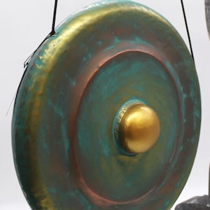 May include: A large, circular gong with a central gold-colored knob. The gong's surface features a blend of teal, bronze, and gold hues, creating a textured, aged appearance. It is suspended by black cords, suggesting it's ready to be played.