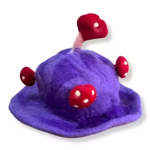 Puede incluir: Un sombrero de fieltro morado caprichoso con tres adornos de setas rojas con lunares blancos. El sombrero tiene un ala ancha y un tallo central de seta.