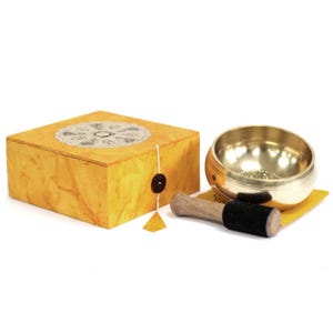 Puede incluir: Un cuenco tibetano dorado con un mazo de madera y una caja amarilla. El cuenco de metal tiene una forma pequeña y redonda. El mazo tiene un mango de madera y una cabeza de fieltro negro. La caja es amarilla con un diseño decorativo.