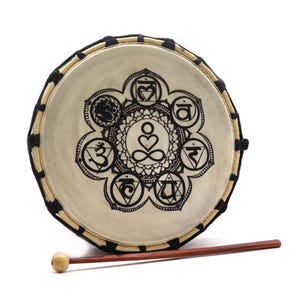 May include: Round drum with a cream-colored drumhead featuring a black chakra symbol design. The drum has a wooden frame and is accompanied by a wooden drumstick with a rounded head. The drum is designed for meditation and spiritual practices.