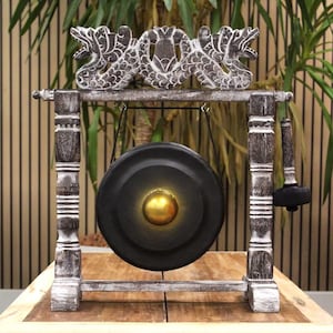 May include: A decorative gong with a black metal disc and a gold center, suspended within a wooden frame. The frame is painted in a distressed white and gray finish, with carved dragon-like figures at the top. A black mallet rests on the side.