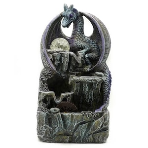 May include: A detailed dragon fountain sculpture. The dragon is perched atop a rocky structure with a glowing orb. The sculpture features shades of grey, blue, and purple, with a waterfall effect. The fountain is designed for indoor or outdoor use.