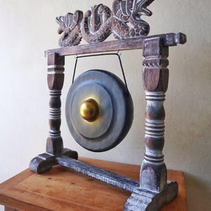 May include: A decorative gong with a wooden frame. The gong is gray with a gold center and is suspended from a frame with carved dragon details. The frame is dark brown with white accents and sits on a wooden surface.
