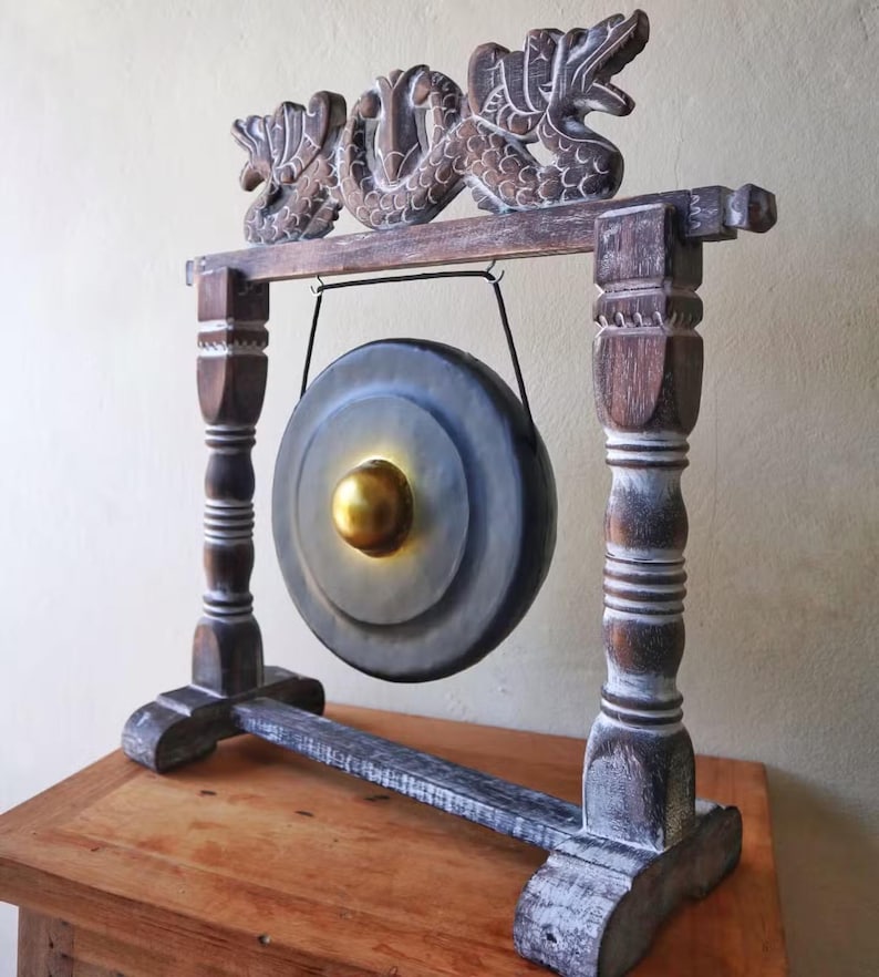 May include: A decorative gong with a wooden frame. The gong is gray with a gold center and is suspended from a frame with carved dragon details. The frame is dark brown with white accents and sits on a wooden surface.