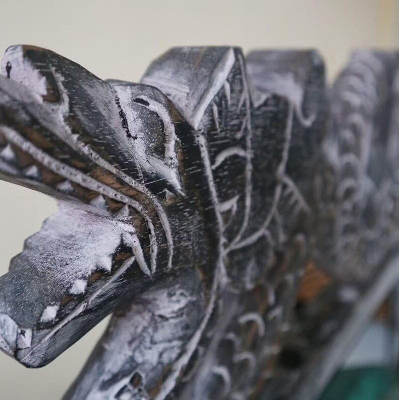 May include: Close-up of a carved wooden sculpture with a dragon-like head. The sculpture is painted in dark gray with white accents, highlighting the intricate details of the carving. The artwork appears to be part of a larger piece.