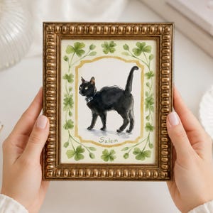May include: A rectangular picture frame with a beaded gold finish containing a watercolour illustration of a black cat wearing a collar. The cat is centred within a border of green clover leaves, and the name Salem is written below the cat.