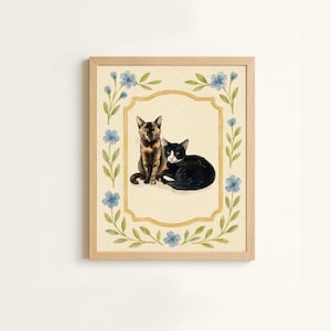 Puede incluir: Una ilustración en acuarela enmarcada que muestra un gato carey sentado junto a un gato blanco y negro tumbado. Los gatos están centrados dentro de un borde dorado, rodeados por una corona decorativa de flores azules y hojas verdes.