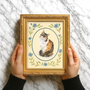 Op de afbeelding: Een ingelijst kattenportret. Het kunstwerk toont een lapjeskat in een blauwe ovaal, omringd door blauwe bloemen en groene bladeren op een crèmekleurige achtergrond. De lijst is goudkleurig.