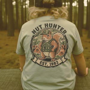 May include: Light blue t-shirt with a graphic of a squirrel wearing a camouflage hat and vest, with the words "NUT HUNTER" and "EST. 1982" in a circular design. The squirrel is holding a nut. The shirt is short-sleeved.