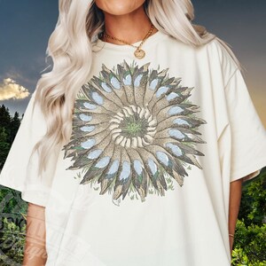 May include: A cream-coloured t-shirt with a circular design of stylised bird feathers in brown, blue, and green. The design is centred on the shirt, creating a symmetrical pattern. The shirt has short sleeves and a relaxed fit.