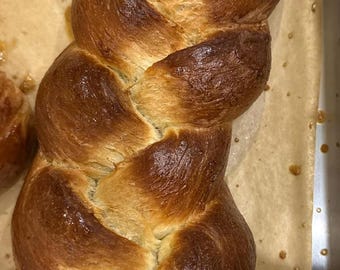 May include: A golden-brown, braided loaf of bread, likely challah, sits on parchment paper in a metal baking pan. The bread has a glossy, baked appearance with a rich, caramelized crust. The texture suggests a soft interior.