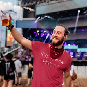 Puede incluir: Una persona con una camiseta roja con el texto "Fetch me an IPA, would ya?" en blanco. La persona sostiene un vaso de cerveza y sonríe.