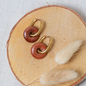 May include: Gold-coloured hoop earrings featuring a circular, reddish-brown stone centre. The earrings are presented on a wooden slice with dried decorative plants.