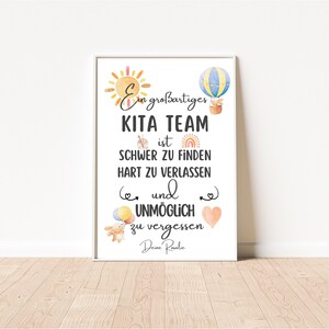 May include: A framed print with the text "Ein gro&szlig;artiges KITA TEAM ist SCHWER ZU FINDEN HART ZU VERLASSEN und UNM&Ouml;GLICH zu vergessen Deine Rovalic." The print features watercolor illustrations of a sun, hot air balloon, rainbow, and hearts.