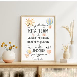 May include: A framed print with the text "Ein gro&szlig;artiges KITA TEAM ist SCHWER ZU FINDEN HART ZU VERLASSEN und UNM&Ouml;GLICH zu vergessen Deine Rosalie." The print has watercolor illustrations of a sun, hot air balloon, rainbow, and other elements.