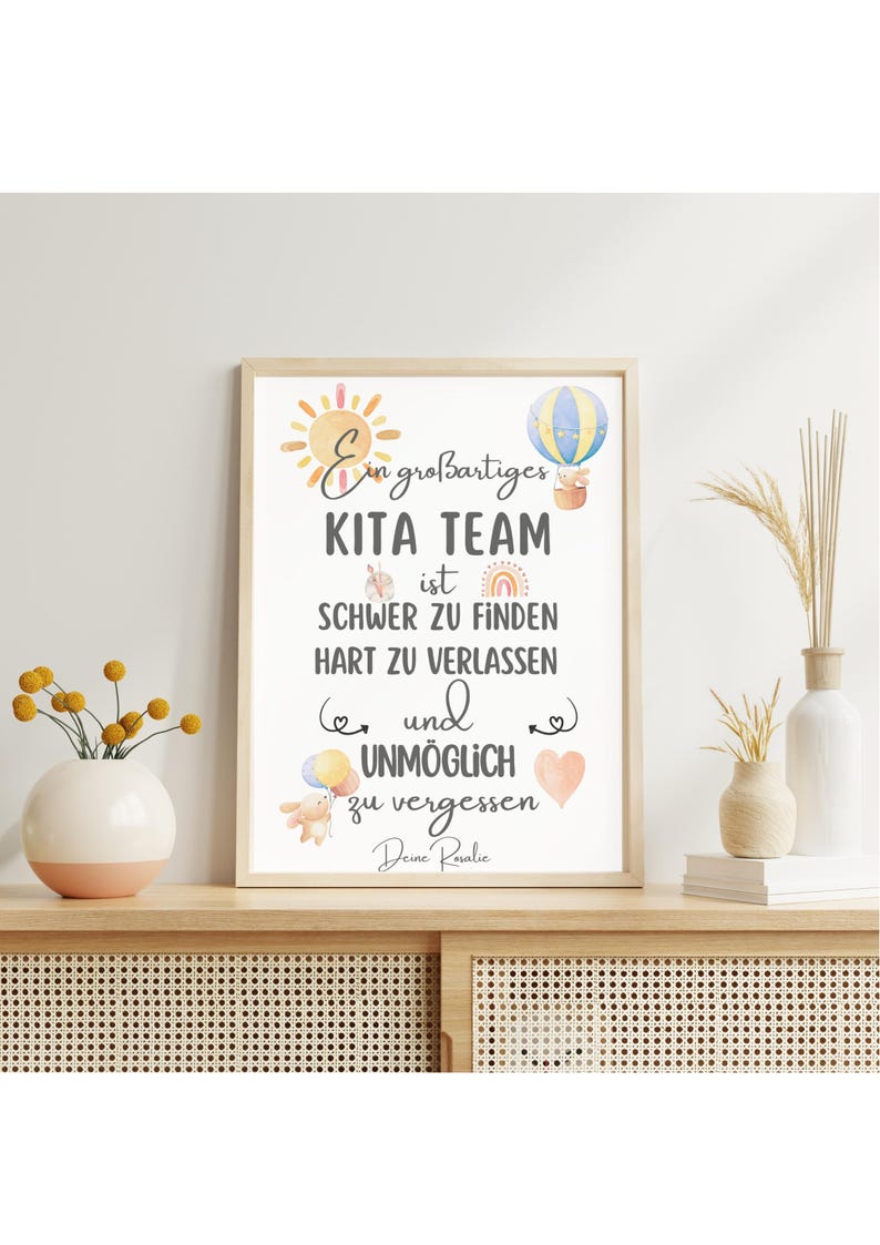 May include: A framed print with the text "Ein gro&szlig;artiges KITA TEAM ist SCHWER ZU FINDEN HART ZU VERLASSEN und UNM&Ouml;GLICH zu vergessen Deine Rosalie." The print has watercolor illustrations of a sun, hot air balloon, rainbow, and other elements.