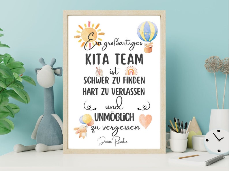 May include: A framed print with a light wood frame features the text "Ein gro&szlig;artiges KITA TEAM ist SCHWER ZU FINDEN HART ZU VERLASSEN und UNM&Ouml;GLICH zu vergessen Deine Rosalie." The print includes watercolor illustrations of a sun, hot air balloon, and a teddy bear.