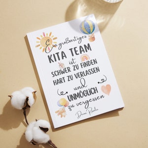 May include: A white notebook with the text "Ein gro&szlig;artiges KITA TEAM ist SCHWER ZU FINDEN HART ZU VERLASSEN und UNM&Ouml;GLICH zu vergessen Deine Rovalic". The cover features watercolor illustrations of a sun, hot air balloon, and other graphics.