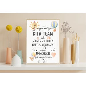 May include: A framed print with the text "Ein gro&szlig;artiges KITA TEAM ist schwer zu finden, hart zu verlassen und unm&ouml;glich zu vergessen." The print is decorated with a sun, hot air balloon, and other illustrations. The print is displayed on a white shelf with vases and decorative floral arrangements.
