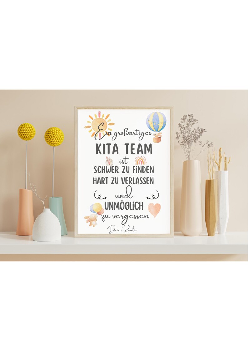 May include: A framed print with the text "Ein gro&szlig;artiges KITA TEAM ist schwer zu finden, hart zu verlassen und unm&ouml;glich zu vergessen." The print is decorated with a sun, hot air balloon, and other illustrations. The print is displayed on a white shelf with vases and decorative floral arrangements.