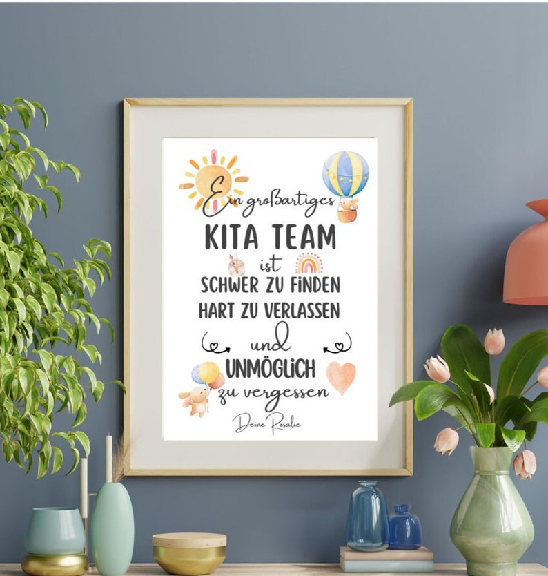 May include: Framed print with the text "Ein gro&szlig;artiges KITA TEAM ist SCHWER ZU FINDEN HART ZU VERLASSEN und UNM&Ouml;GLICH zu vergessen Deine Roralie." The print features watercolor illustrations of a sun, hot air balloon, rainbow, and teddy bear.