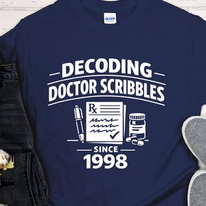 Op de afbeelding: Marineblauw t-shirt met de witte tekst "DECODING DOCTOR SCRIBBLES SINCE 1998". Het ontwerp bevat een receptsymbool, een pen, een pillenfles en een vinkje. De woorden "Custom year available" zijn ook aanwezig.