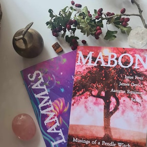 May include: Two magazines, "Lammas" and "Mabon," are displayed with a bronze apple, a pink stone, a clear crystal, and a sprig of berries. The "Mabon" magazine features a tree with pink leaves. The "Lammas" magazine has a purple and blue design.