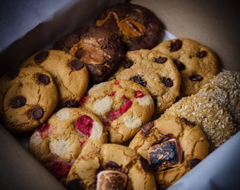 May include: A selection of freshly baked cookies in a cardboard box. The cookies include chocolate chip, raisin, raspberry, and marshmallow flavours. The cookies are arranged in a box with white paper lining.