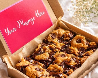 May include: A box of chocolate chip cookie bars with a pink card that reads "Your Message Here". The bars are cut into squares and sit on parchment paper inside a brown cardboard box. The background is a white sheet.