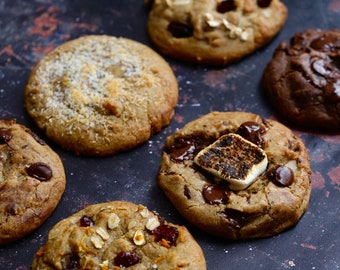 May include: An assortment of freshly baked cookies on a dark surface. The cookies vary in size and toppings, including chocolate chips, oats, cranberries, and a toasted marshmallow. The cookies are golden brown and appear soft.