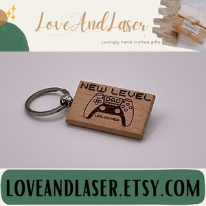 Könnte beinhalten: Holzschlüsselanhänger mit einem lasergravierten Design eines Videospielcontrollers mit dem Text "NEW LEVEL DAD UNLOCKED".