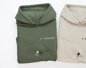 May include: Two hooded sweatshirts, one green and one beige, with the same text embroidered on the front: "X.MMXXIII". The green sweatshirt has a white heart with the letter "I" embroidered on the left sleeve, and the beige sweatshirt has a black heart with the letter "M" embroidered on the left sleeve.