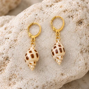 Peut inclure: Une paire de boucles d'oreilles créoles dorées avec des petits coquillages naturels en spirale en guise de breloques. Chaque coquillage est de couleur crème avec un motif distinct de taches rectangulaires brunes, suspendu à un anneau en métal doré.